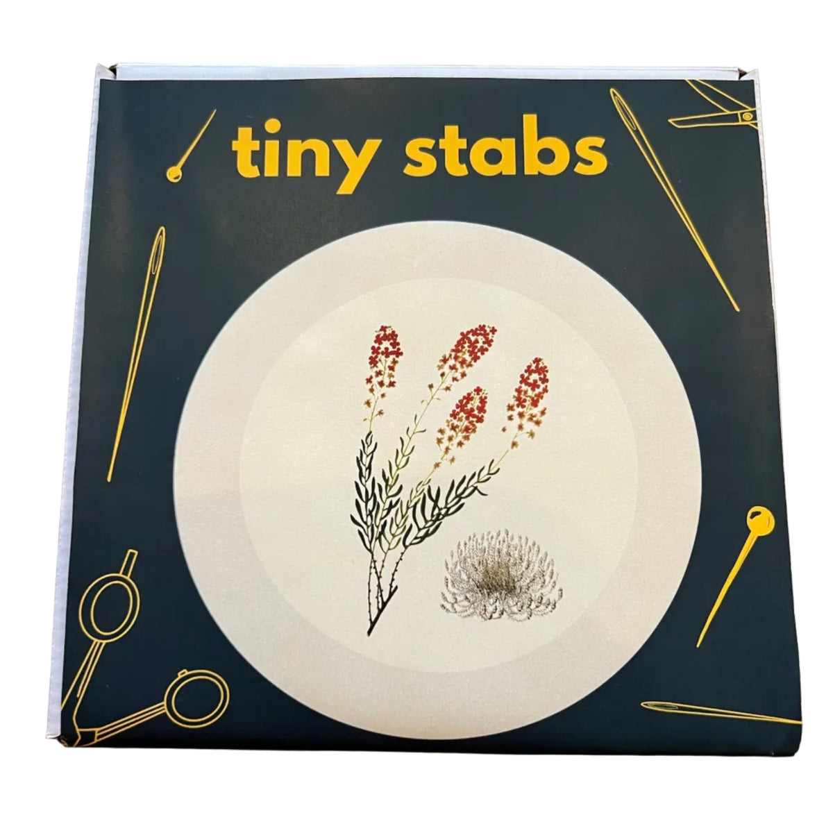 A square product box for a floral "Tiny Stabs" embroidery kit. The box has a dark forest green background with yellow text and yellow line drawings of sewing tools, featuring a central white circular illustration with red and green floral details. 
