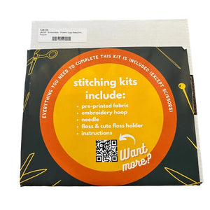 A square product box for an embroidery kit, photographed from above on a wooden surface. The box is dark forest green with a large orange circular label reading 'stitching kits include:' and listing contents such as pre-printed fabric, embroidery hoop, needle, and instructions.