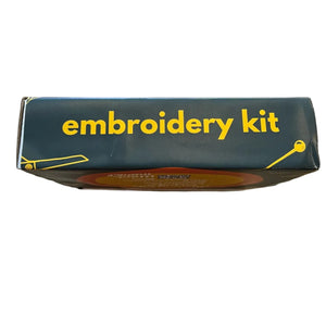 A dark forest green cardboard box labeled 'embroidery kit' in yellow text, photographed from above on a worn wooden tabletop.