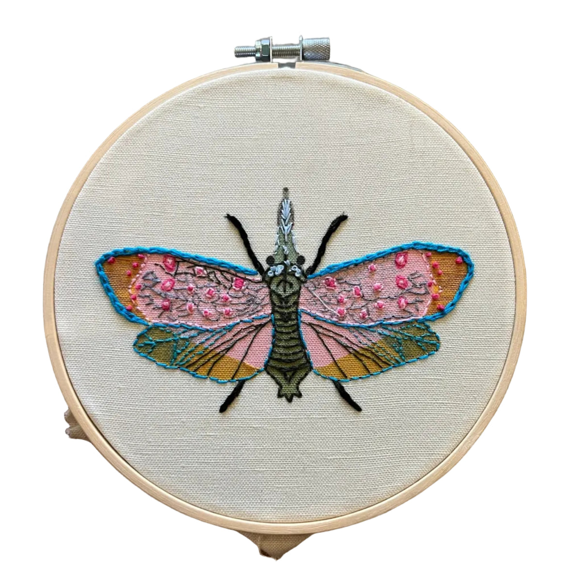 A circular embroidery hoop holding cream fabric with a detailed embroidered moth design. The moth has pink wings with small embellishments, blue outlines, and a dark green body.