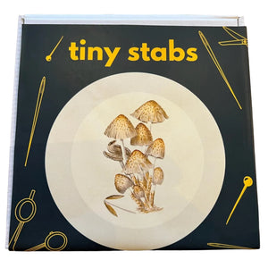 A square product box titled 'tiny stabs' in yellow text, featuring a dark forest green cover with a large white plate illustration and yellow needle and scissor graphics, photographed on a wooden tabletop.