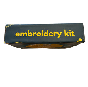 Top-down view of a dark forest green cardboard box labeled 'embroidery kit' in yellow text, placed on a worn wooden tabletop.