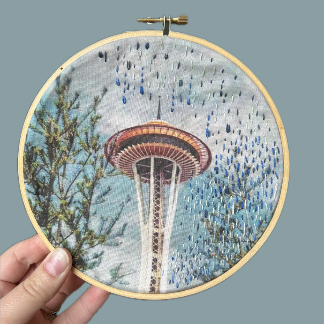 Embroidery hoop with a fabric depiction of the Space Needle in Seattle, held against a gray background.