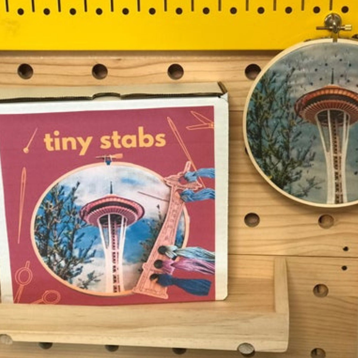 Embroidery hoop with Space Needle design next to a boxed embroidery kit titled 'tiny stabs' on a wooden pegboard.