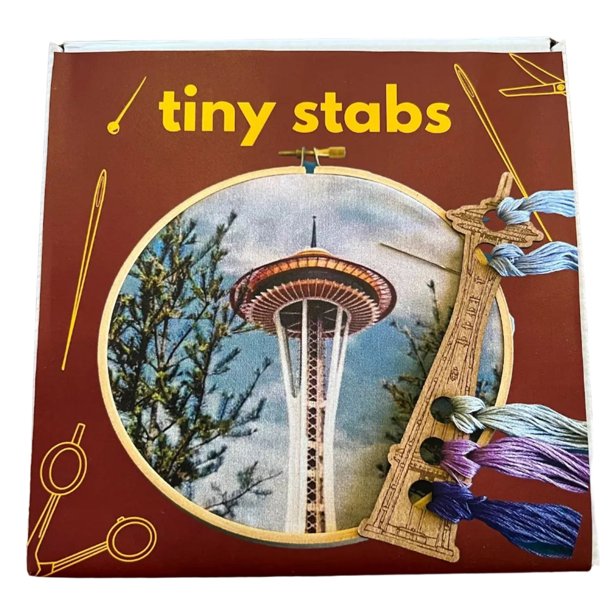 A boxed embroidery kit labeled 'Tiny Stabs'. The box is dark red with yellow text and illustrations, plus a circular photo of the Seattle Space Needle and decorative embroidery floss holder on the right.
