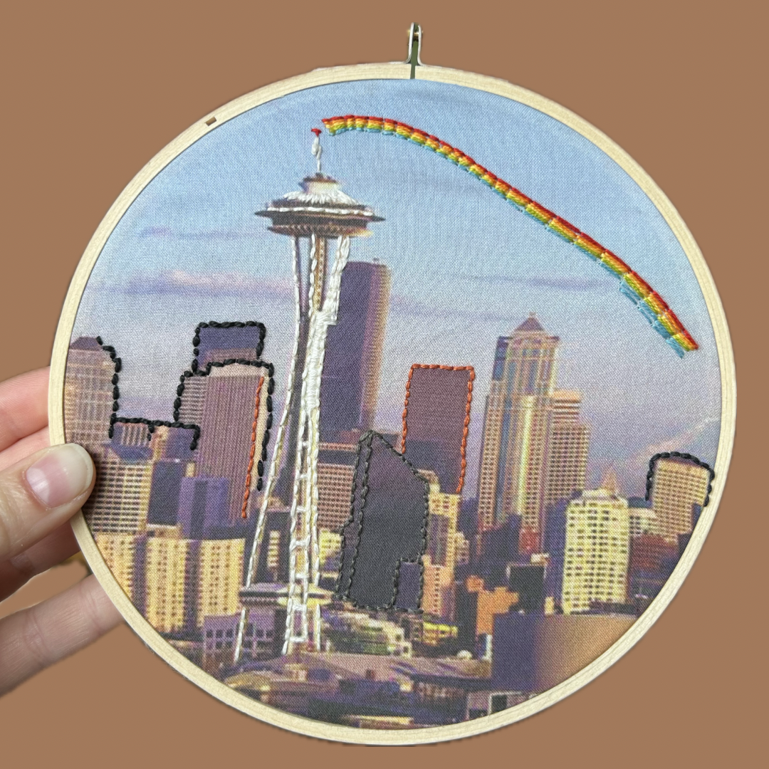 Embroidery hoop with a cityscape including the Space Needle and rainbow on a brown background. 