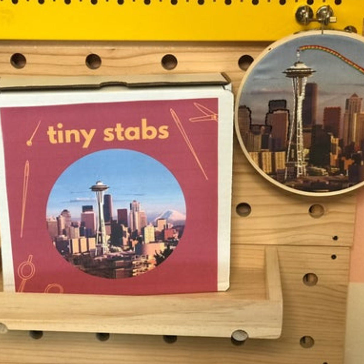 Product display with 'tiny stabs' packaging and an embroidery hoop featuring a Seattle cityscape on a pegboard.