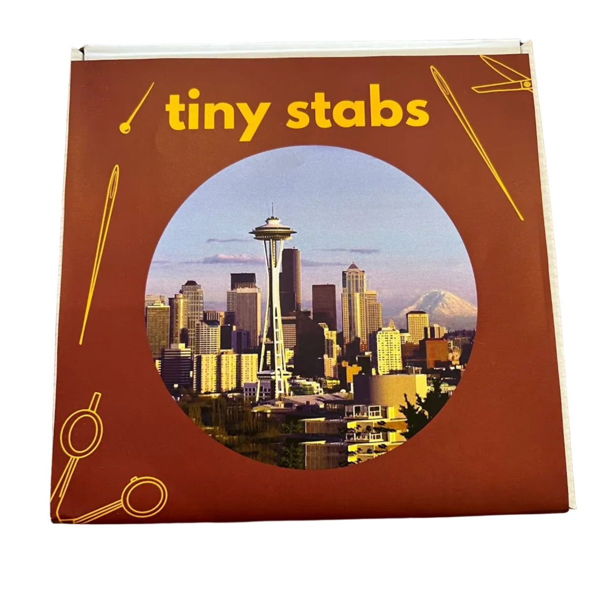A square product box for 'Tiny Stabs' souvenir Seattle embroidery kit. The box features a terracotta red background with yellow text and line-art illustrations of sewing tools, with a circular cutout showing a photo of the Seattle skyline featuring the Space Needle.