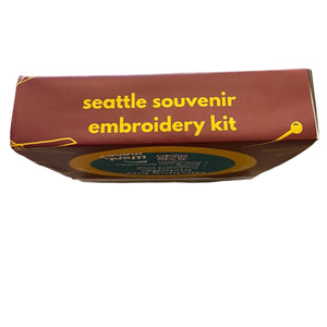 A top-down view of a 'Seattle Souvenir Embroidery Kit' box on a worn wooden tabletop. The box is dark reddish-brown with yellow text and a circular teal and yellow logo.