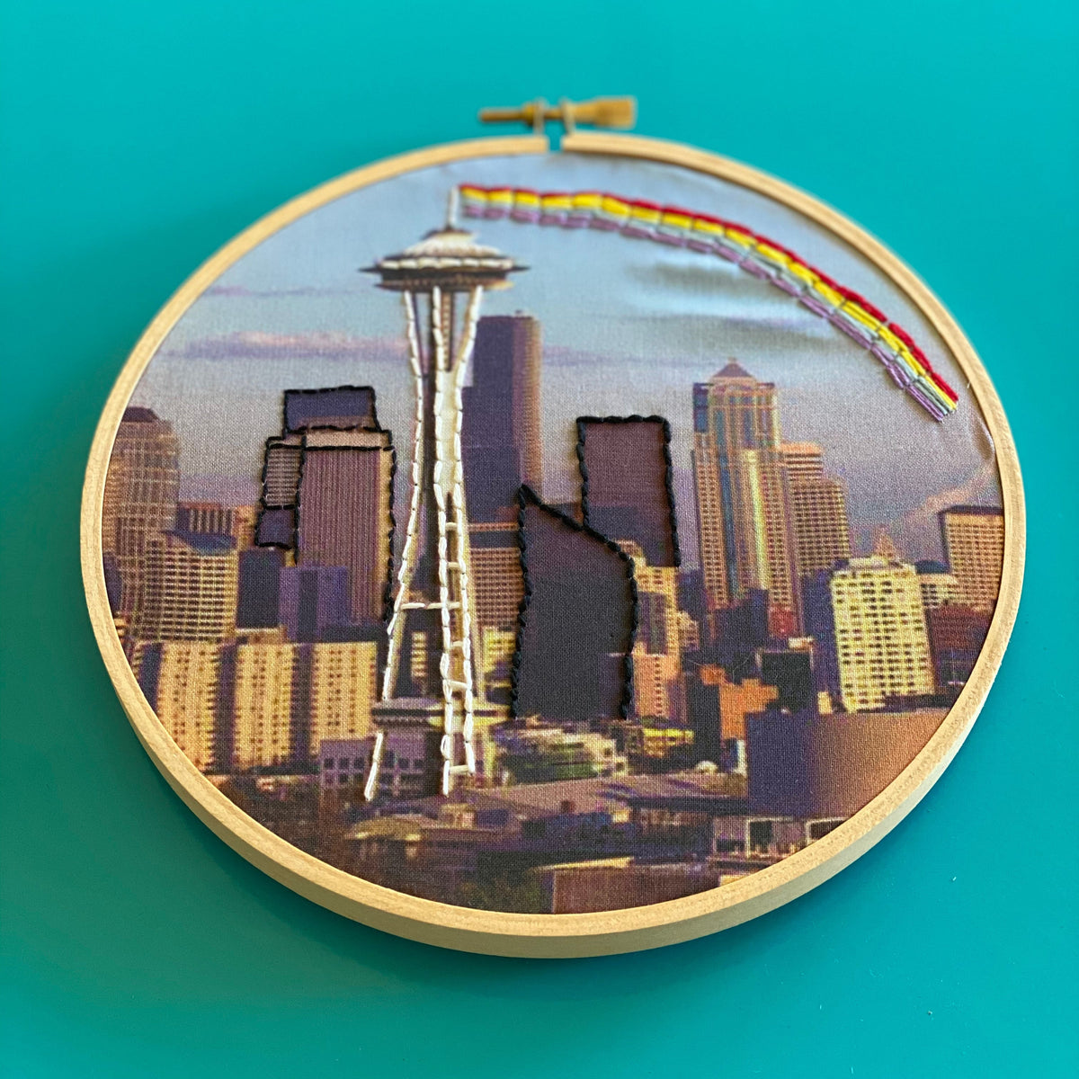 Embroidery hoop with a cityscape including the Space Needle on a teal background. 