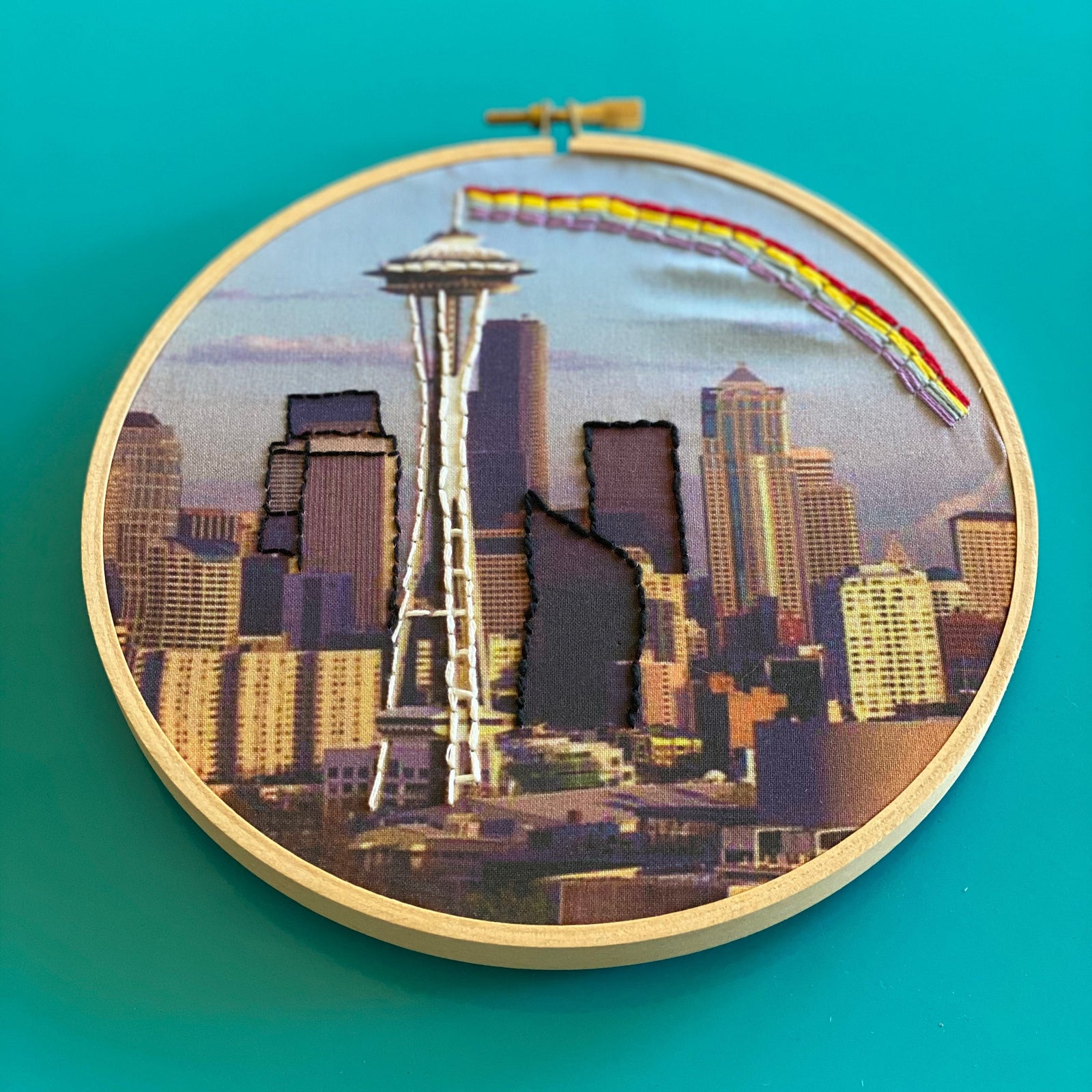 Embroidery hoop with a cityscape including the Space Needle on a teal background. 
