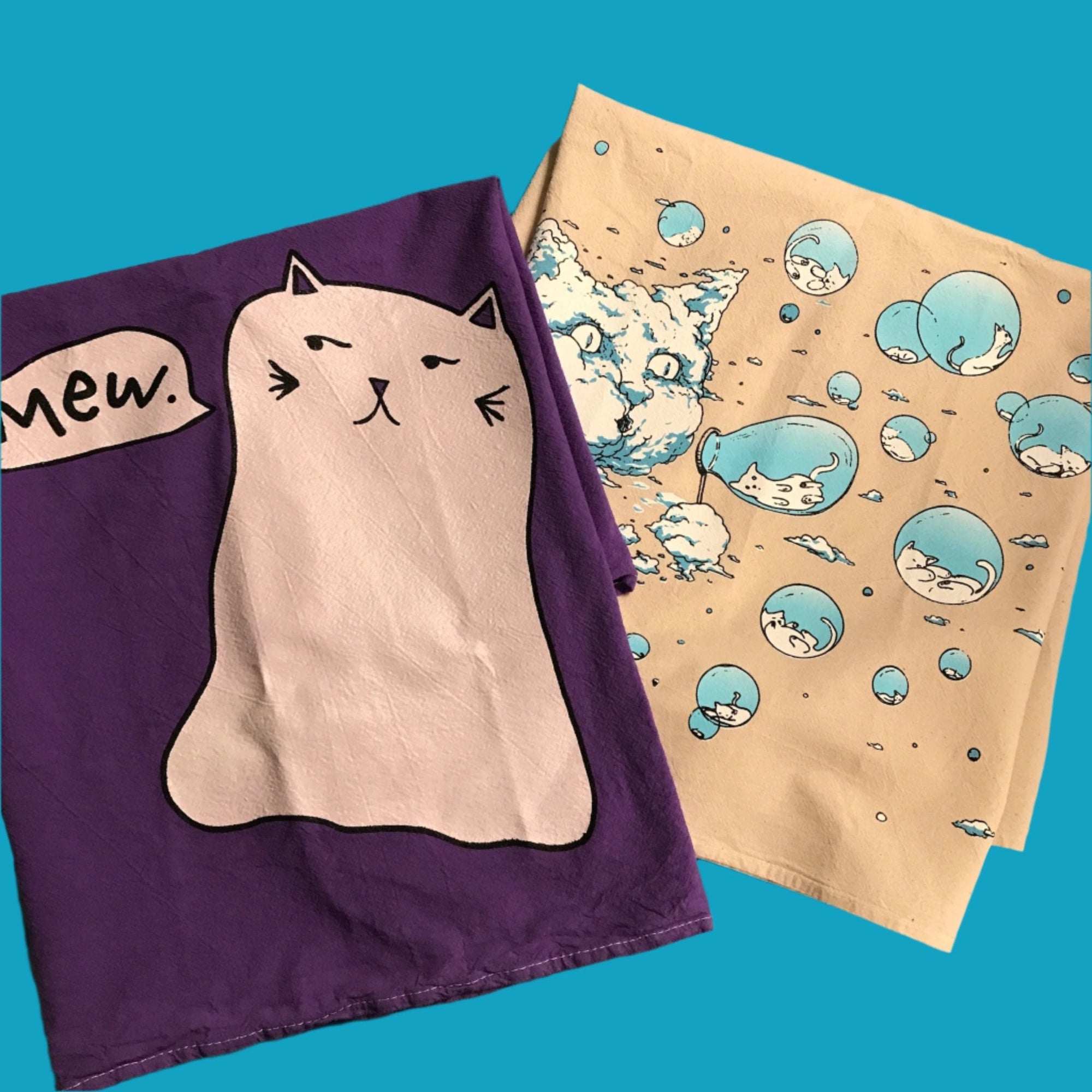 Two kitchen towels are folded and sitting on a teal background. On the bottom there is the Bubble Cat design with a Cloud Cat blowing bubbles and inside the bubbles are sleeping kittens. This is screenprinted in blue and white on a natural color kitchen towel. On top is a large white Ghost Kitty that has a word balloon saying MEW. It is printed in black and white on a dark purple towel.
