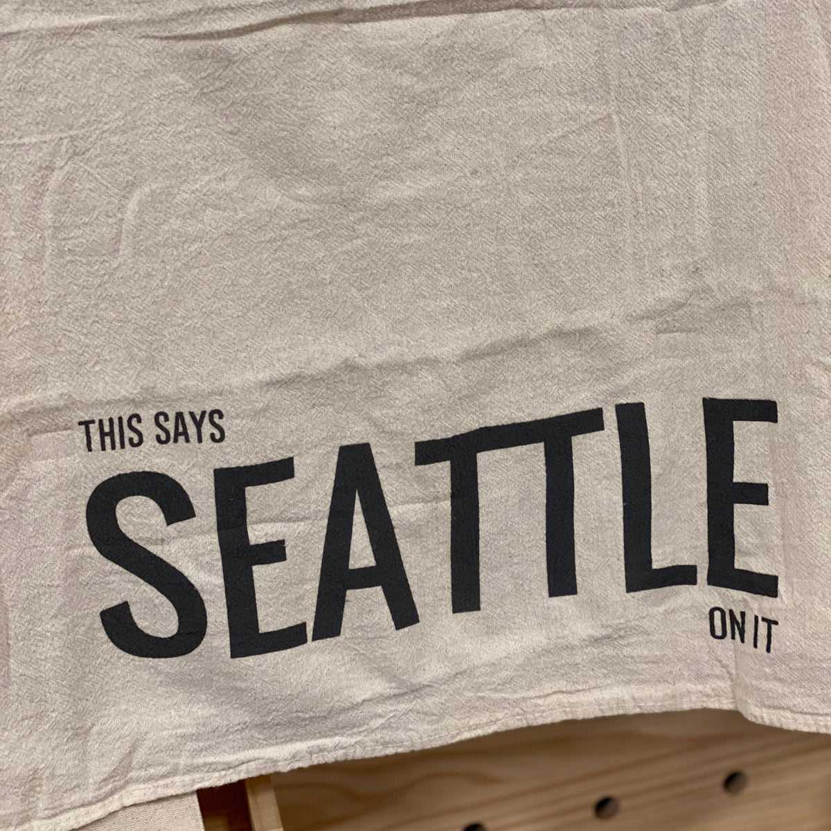 This natural color kitchen towel has "This Says Seattle On It" in dark gray ink. It is hanging in front of a wooden pegboard wall. 