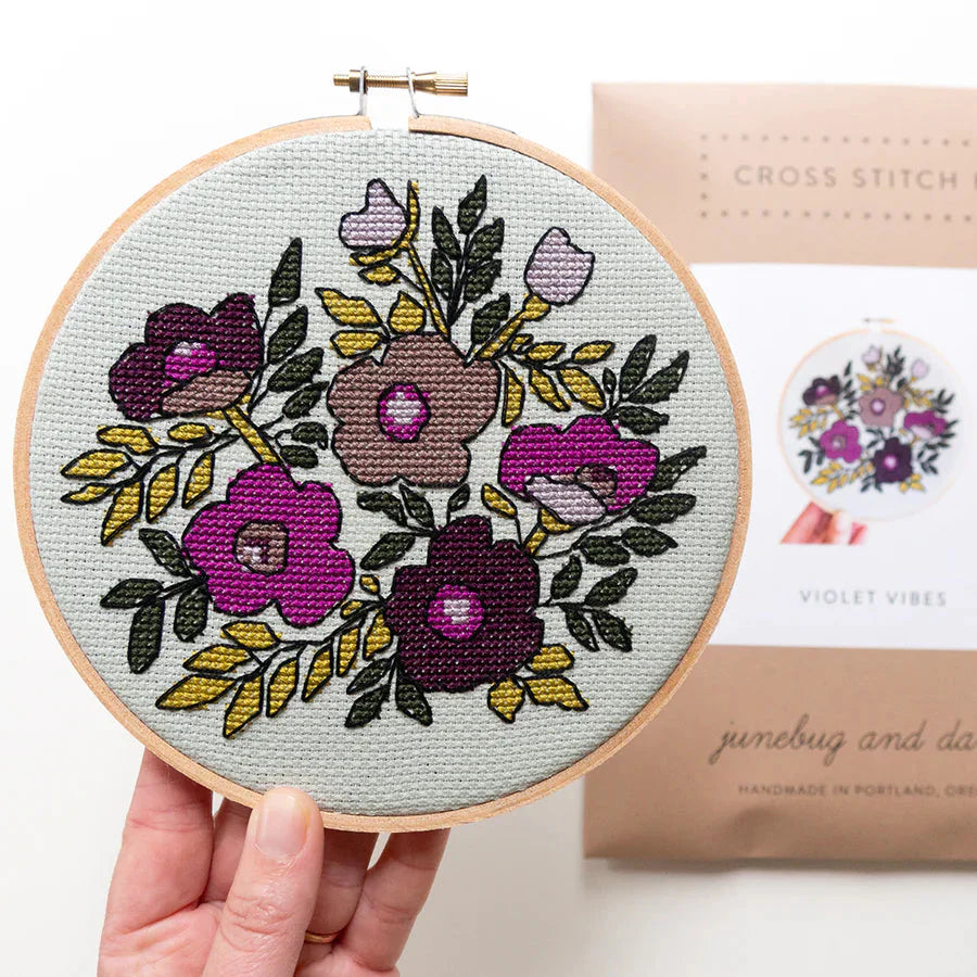 A stitched up sample of the Violet Vibes cross stitch pattern is being held up by a human hand. The packaging for the kit is blurry in the background. 