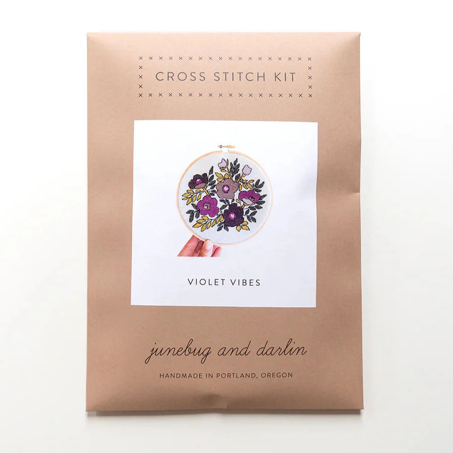The packaging for the Violet Vibes cross stitch kit is a kraft brown envelope that says "Cross stitch kit" at the top. It has a picture of the finished kit on the front. 