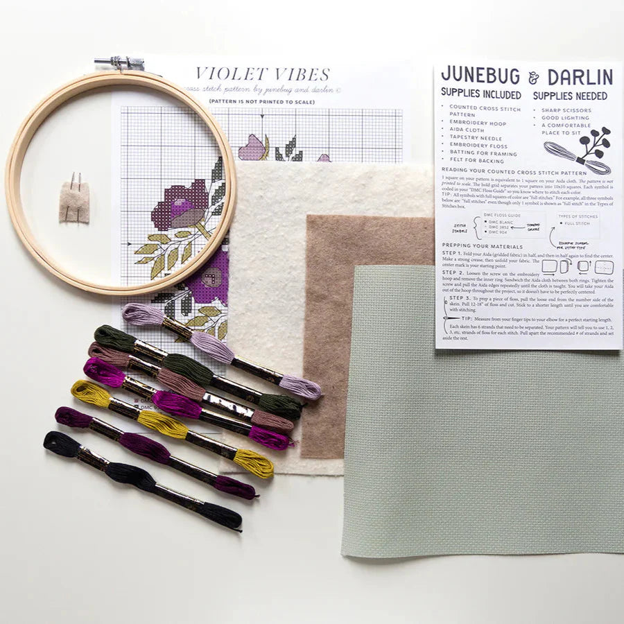 All of the supplies you get in the Violet Vibes cross stitch kit laid out and photographed from above. You get an embroidery hoop, needles, embroidery floss, aida fabric, backing felt, pattern and hand illustrated instructions. 