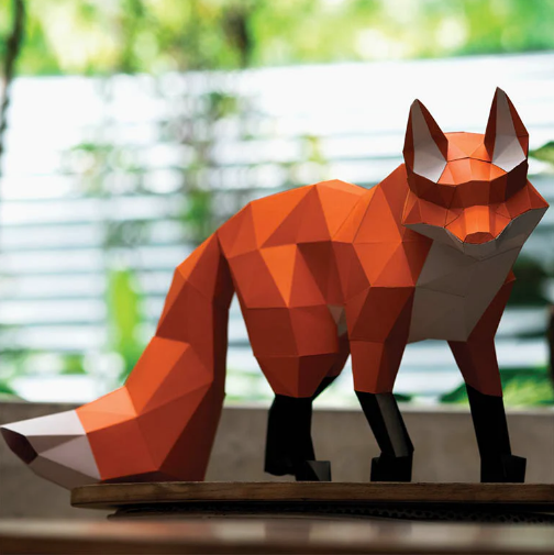 A finished example of a walking fox paper craft kit. It is on a table and shown in front of a blurry, sunny background. 