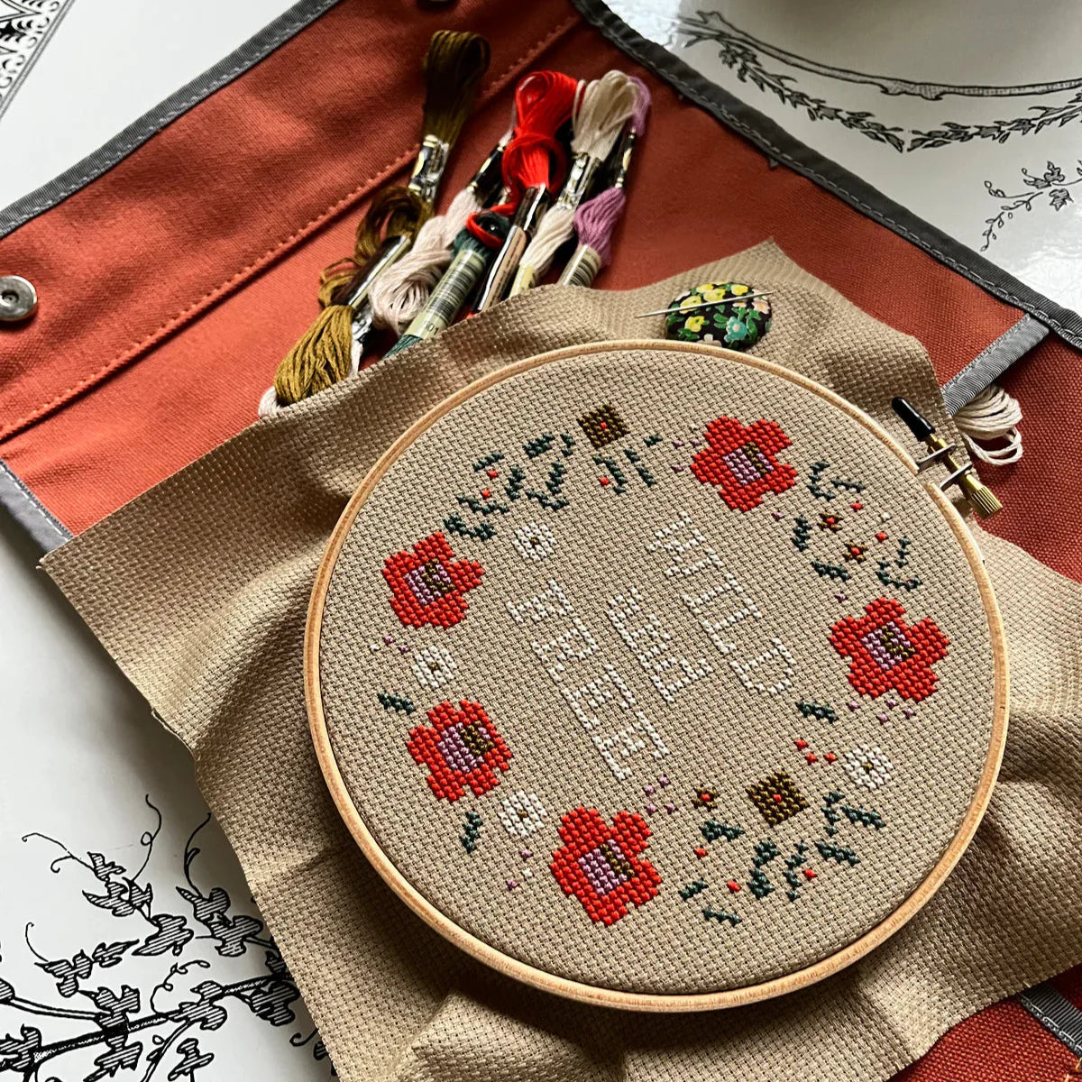 A stitching up example of the Wild and Free pattern in progress. The aida fabric is set in the hoop and embroidery floss is set to the side. A needle on a needle minder is on the aida fabric. 