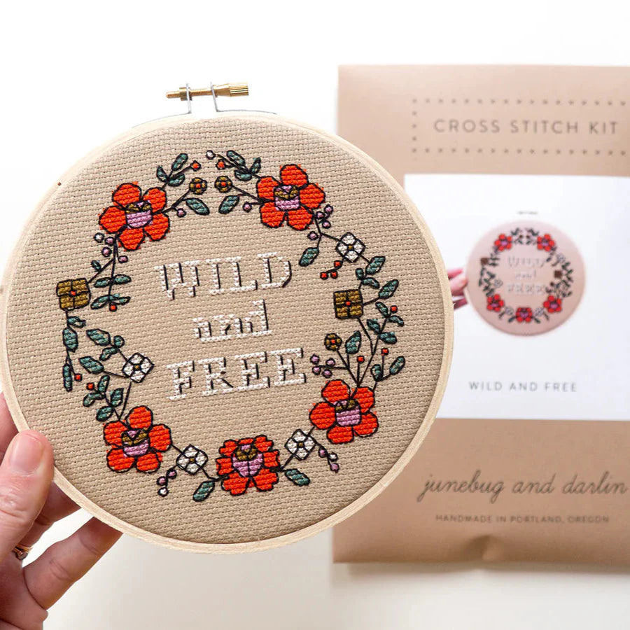 A stitched up example of the Wild and Free cross stitch kit being held up by a human hand. The packaging for the kit is blurry in the background. The kit has the words in the center of a circle of brightly colored flowers.