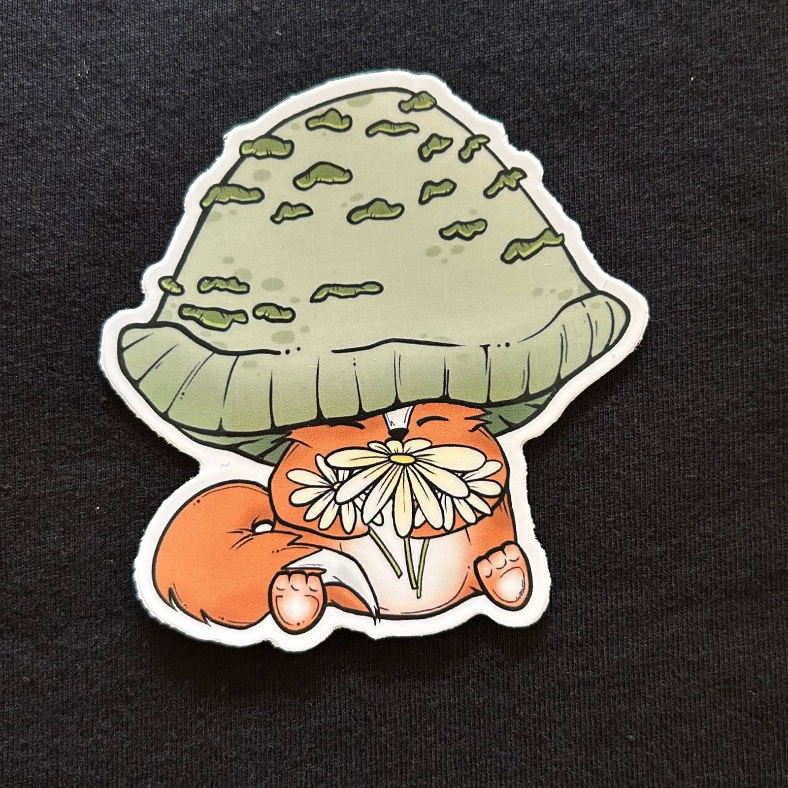 A stocker depicting a fox wearing a green mushroom as a hat holding a bouquet of daisies. 