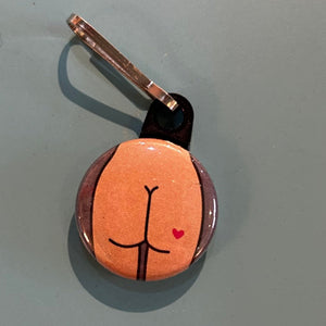 Small round zipper pull with a butt design with a heart tattoo on a light gray background. 