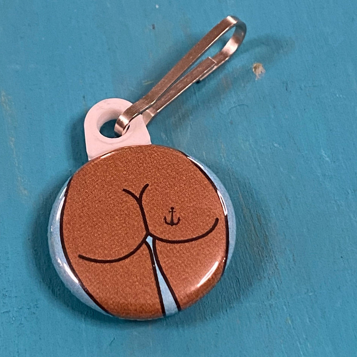 Round zipper pull of a round butt with an anchor tattoo on a blue background. 