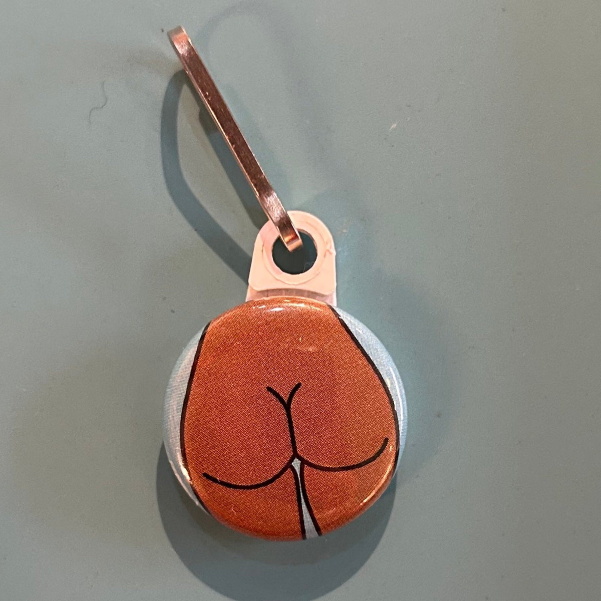 Small round zipper pull with a round butt design on a gray background.