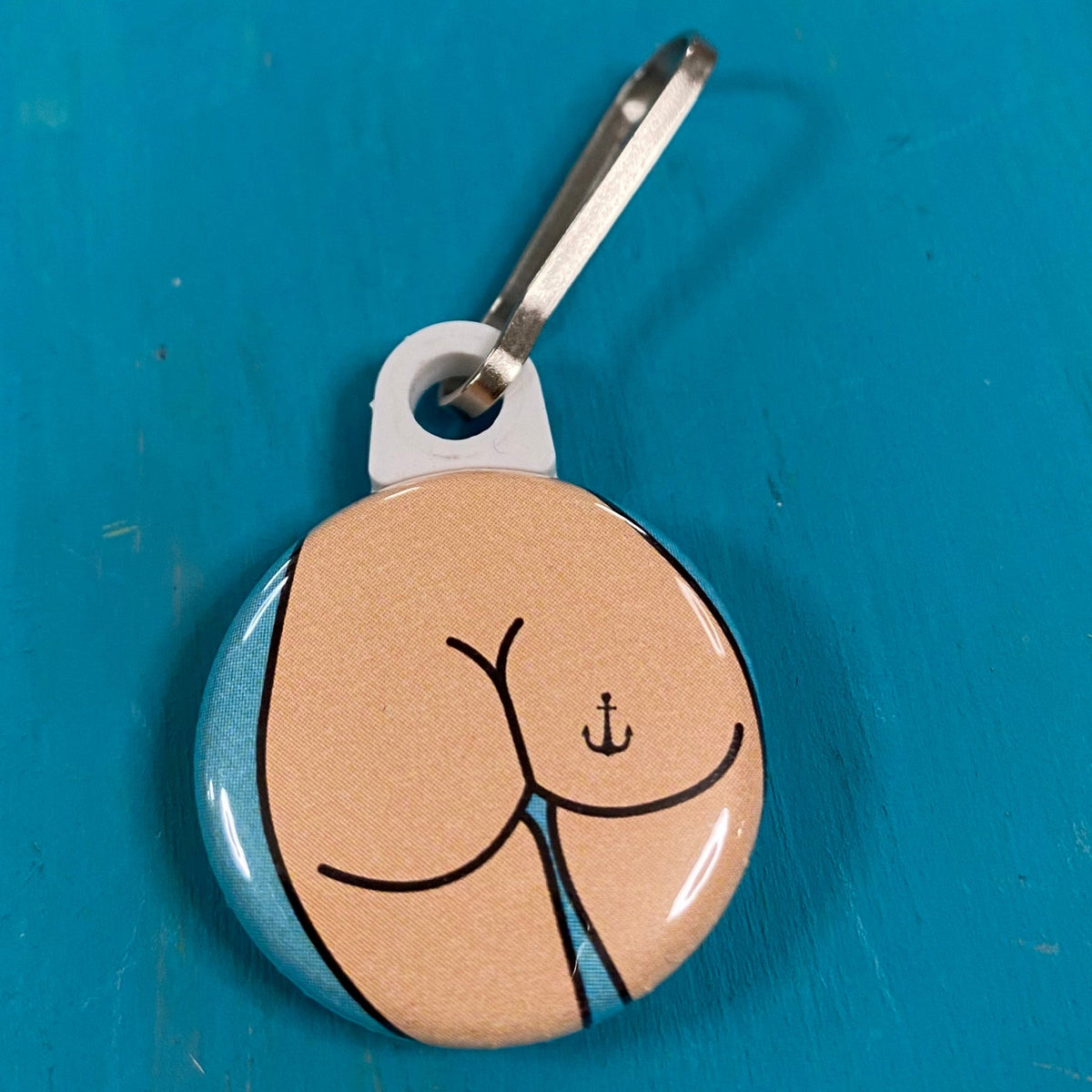 Round zipper pull with a butt design with an anchor tattoo on a blue surface. 