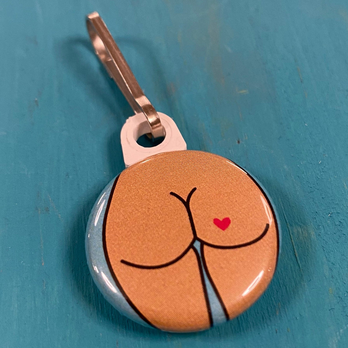 Butt-themed zipper pull on a blue surface. It has a heart-shaped tattoo.