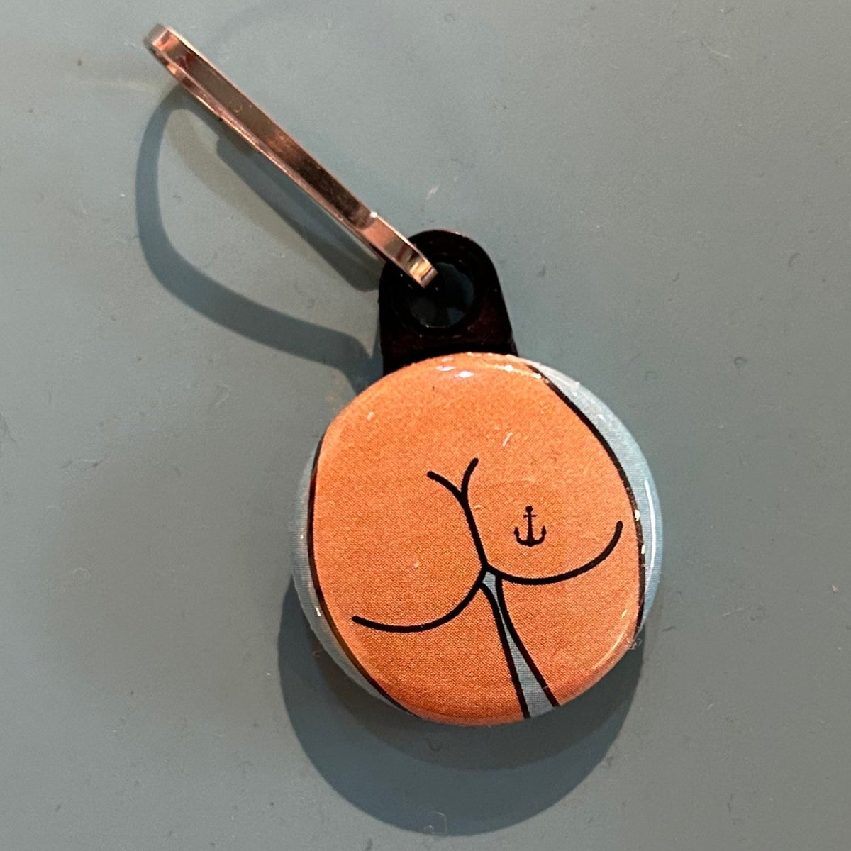zipper pull with a butt design with an anchor tattoo on a gray background. 