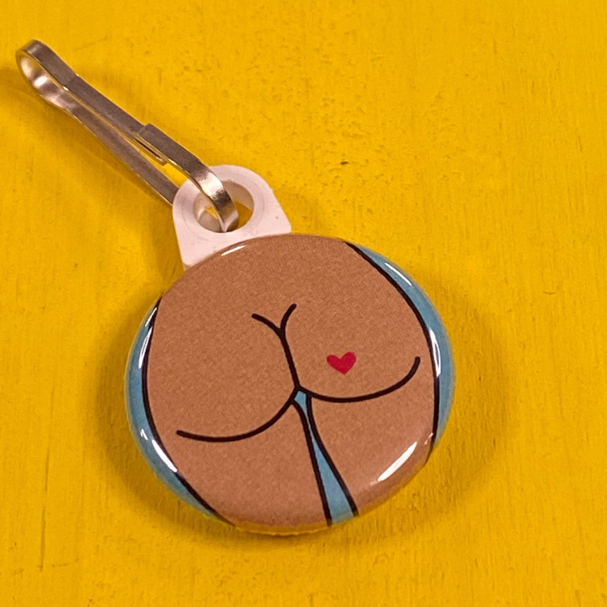 Round zipper pull with a round butt and a heart tattoo on a yellow surface. 