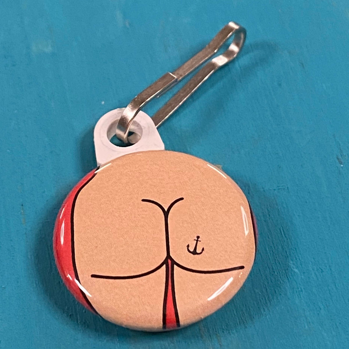 Round zipper pull with a butt design with an anchor tattoo on a blue background. 
