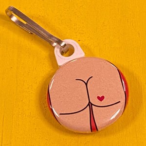 Round zipper pull with a round butt design with a heart tattoo on a yellow surface. 