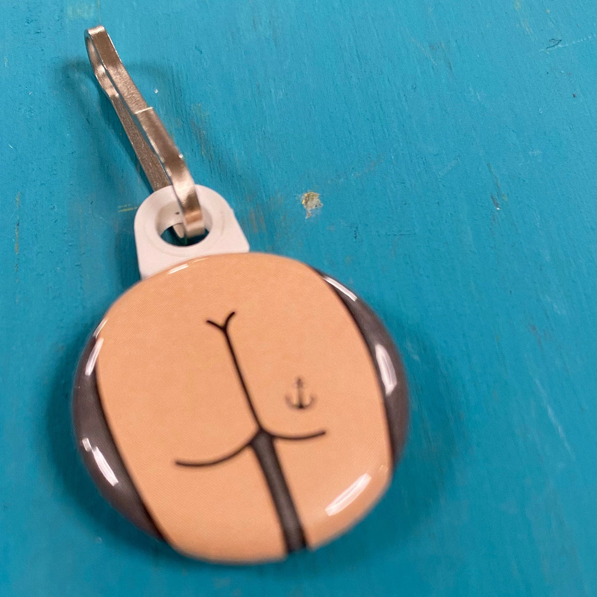 Round zipper pull with a butt design with an anchor tattoo on a blue background. 