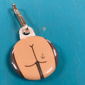 Round zipper pull with a butt design with an anchor tattoo on a blue background. 