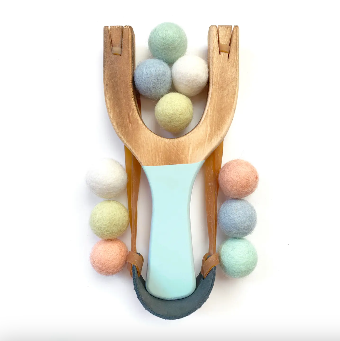 A wooden slingshot toy with a pale blue handle, arranged on a white background with pastel felt balls around and inside it.