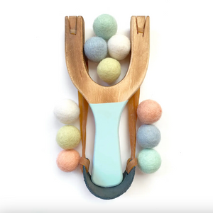 A wooden slingshot toy with a pale blue handle, arranged on a white background with pastel felt balls around and inside it.