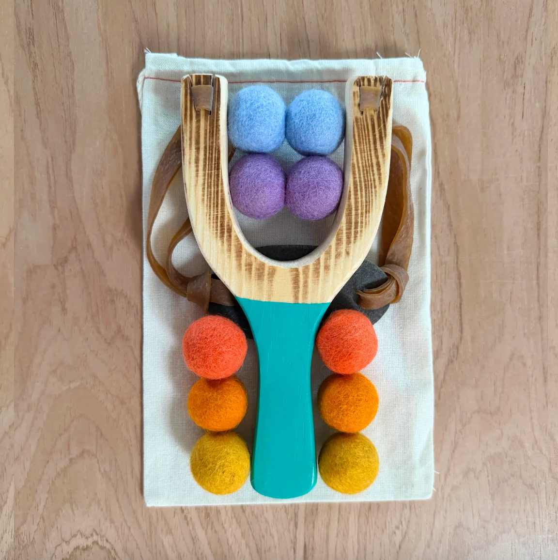 A wooden slingshot toy with a turquoise handle, brown straps, and colorful felt balls in blue, purple, orange, yellow, and brown, displayed on a white fabric mat over a light wood background.