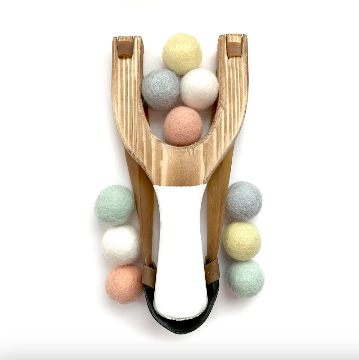 A wooden toy slingshot with a white handle, surrounded with pastel felt balls, on a clean white background.