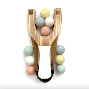 A wooden toy slingshot with a white handle, surrounded with pastel felt balls, on a clean white background.