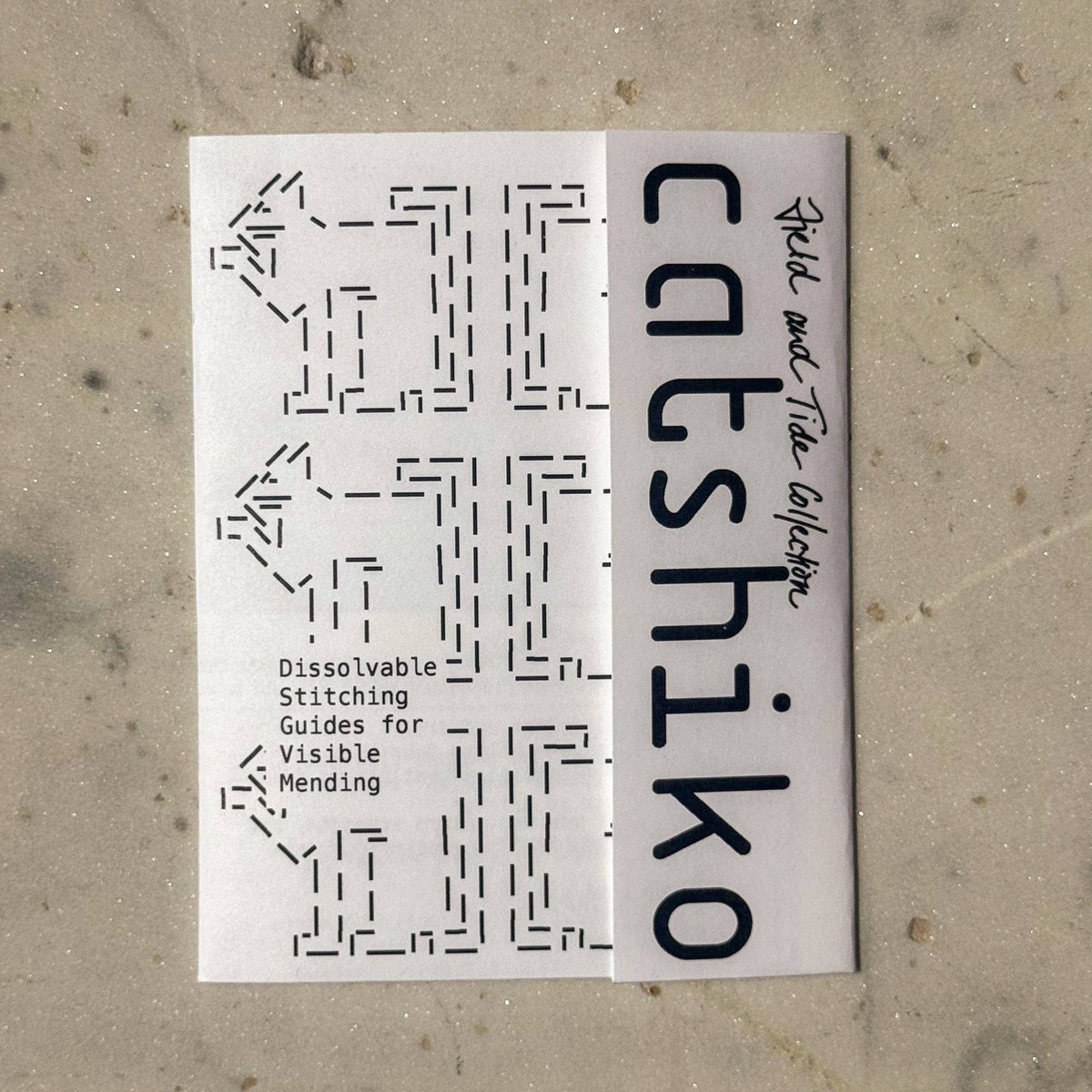 A white rectangular booklet or card titled 'catshiiko' with black text and dashed stitch-pattern graphics, placed on a light beige textured surface.