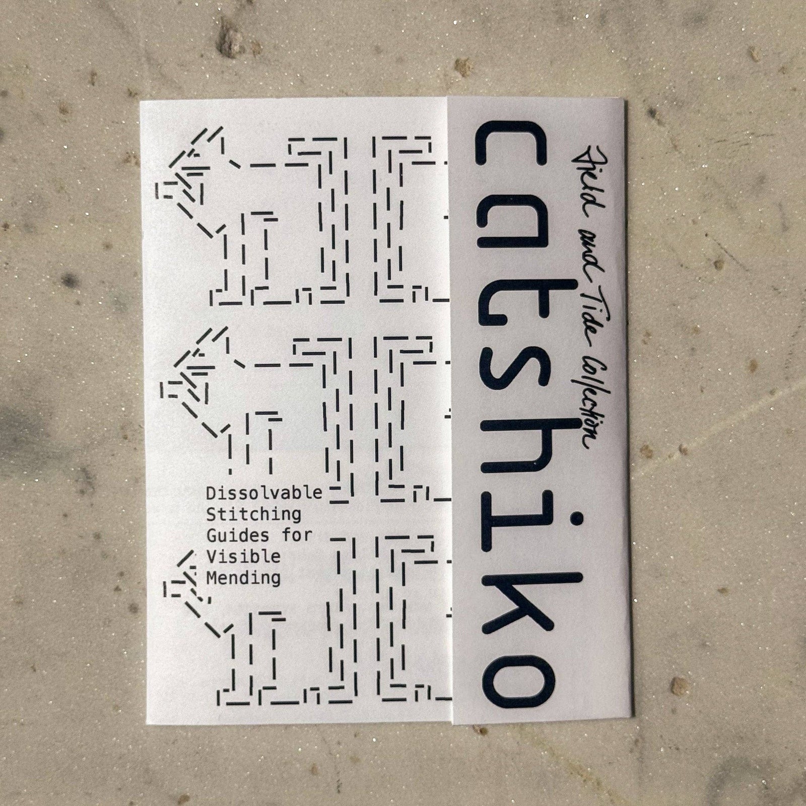 A white rectangular booklet or card titled 'catshiiko' with black text and dashed stitch-pattern graphics, placed on a light beige textured surface.