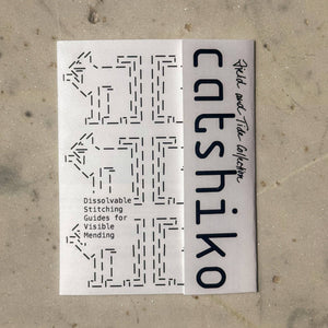 A white rectangular booklet or card titled 'catshiiko' with black text and dashed stitch-pattern graphics, placed on a light beige textured surface.