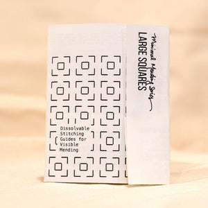 A white booklet titled 'LARGE SQUARES Dissolvable Stitching Guides for Visible Mending' standing upright on a warm beige surface.