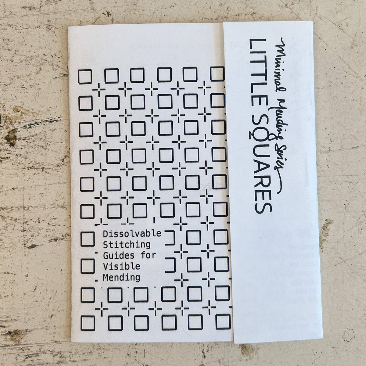 A white booklet  titled 'LITTLE SQUARES' with 'Minimalist Stitching' written on the side, placed on a textured beige surface. The front shows a grid of black square-and-cross stitch patterns with text reading 'Dissolvable Stitching Guides for Visible Mending.