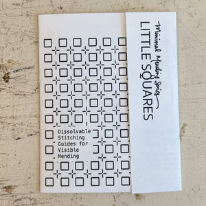 A white booklet  titled 'LITTLE SQUARES' with 'Minimalist Stitching' written on the side, placed on a textured beige surface. The front shows a grid of black square-and-cross stitch patterns with text reading 'Dissolvable Stitching Guides for Visible Mending.