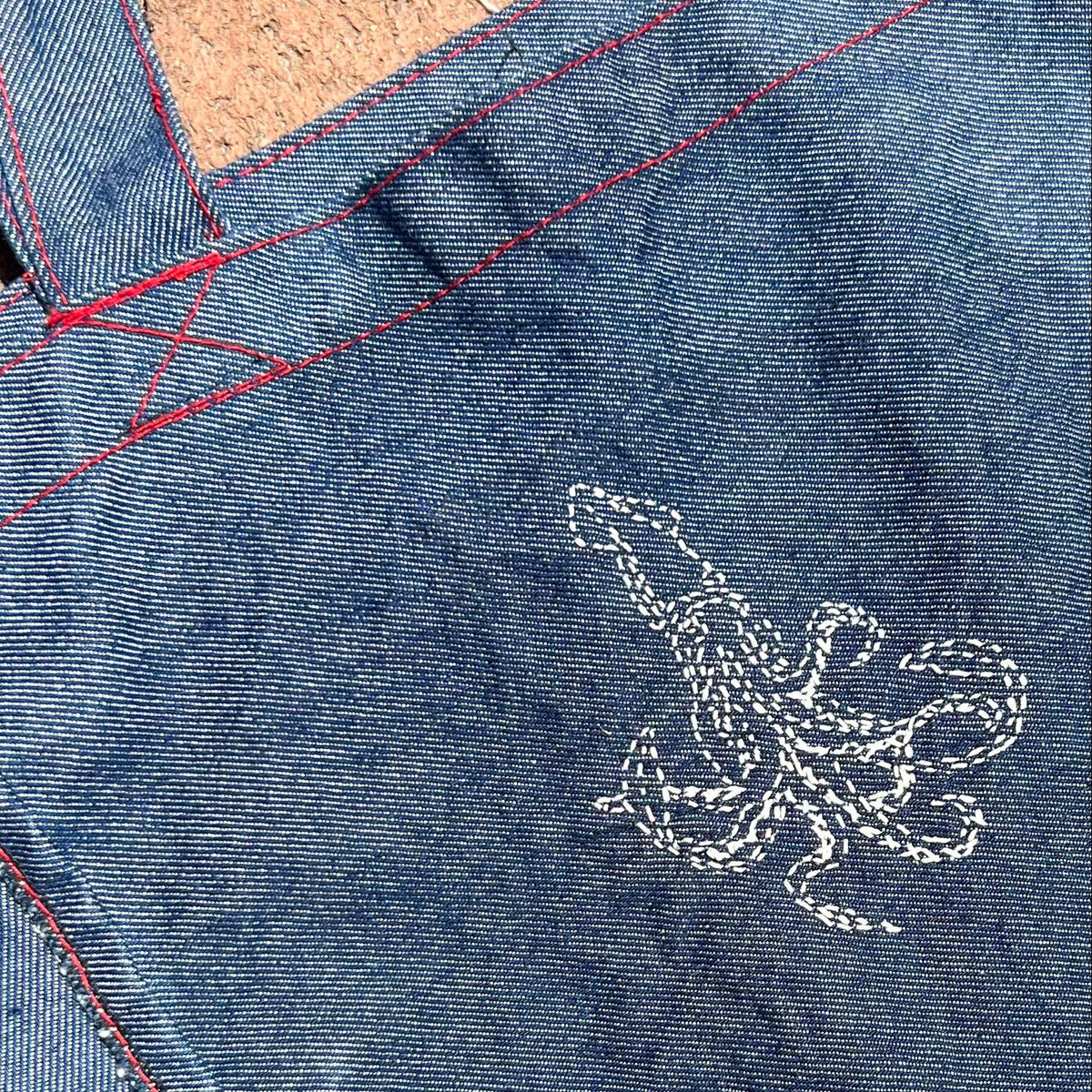 Close-up of a blue denim tote bag showing the fabric texture, red contrast stitching, and a white embroidered squid design.