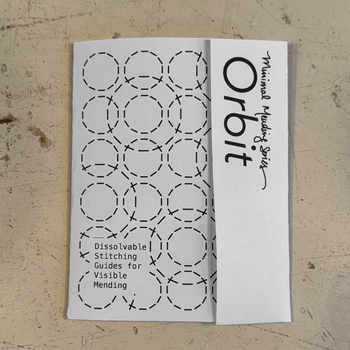 A white paper stitch guide booklet titled 'Minimal Mending Series: Orbit' featuring black printed dashed circle stitching guides and text, placed on a textured beige surface.