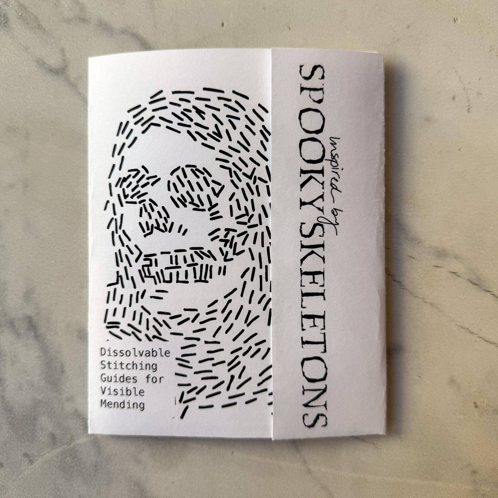 A folded white booklet  titled 'Spooky Skeletons' with black text and a black line-art skull illustration, photographed on a light marble-like surface.