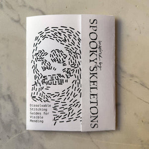A folded white booklet  titled 'Spooky Skeletons' with black text and a black line-art skull illustration, photographed on a light marble-like surface.